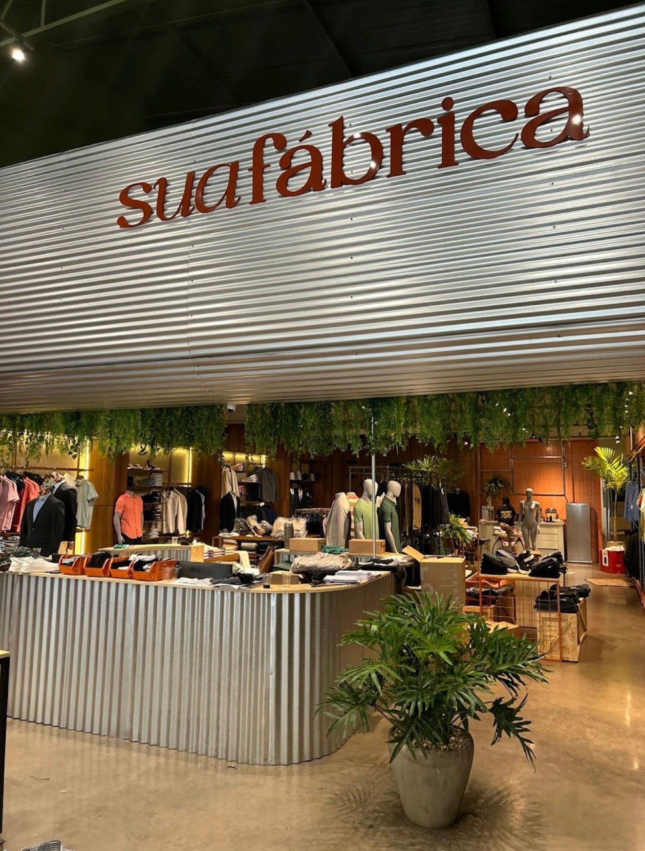 Fachada e interior da loja Sua Fábrica, com o nome da marca na estrutura metálica e araras de roupas